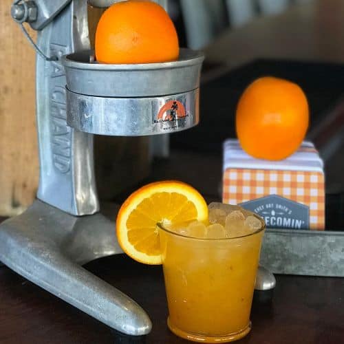 A shiny metal juicer with a fresh orange, a tall glass of iced orange drink, and whole oranges sit on the table, capturing the zesty spirit of Disney Springs’ Florida-Inspired Menu—an ideal way to toast National Florida Day at Walt Disney World’s vibrant dining and entertainment district.