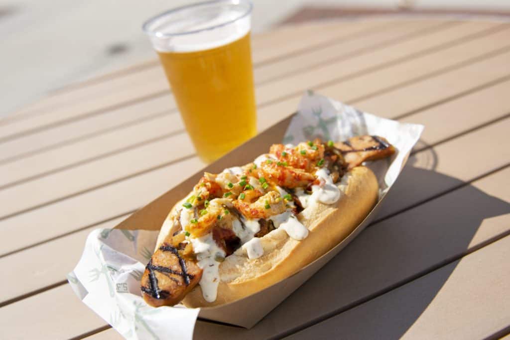 The Smoked Crawdog—grilled sausage loaded with vibrant toppings and sauce in a fresh bun—is paired with a chilled glass of beer, all set on a rustic wooden table outdoors at Disney Springs. This festive dish delivers the bold, celebratory flavors of Mardi Gras straight to your culinary adventure at Walt Disney World Resort.