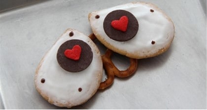 Adorable owl-faced cookies with heart decorations, frosted in white icing and perched on a pretzel base, displayed on a themed treat tray—ready to delight visitors at your favorite theme park’s Valentine’s Day celebration.
