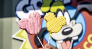 Two vibrant Mickey Mouse-shaped treats on sticks—one pink, one yellow—held up in front of a playful Disney-themed mural. These colorful snacks are the latest must-try delights now available at Disney Springs.