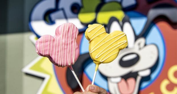 Two vibrant Mickey Mouse-shaped treats on sticks—one pink, one yellow—held up in front of a playful Disney-themed mural. These colorful snacks are the latest must-try delights now available at Disney Springs.