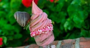 Strawberry soft serve served in a glass at Disney Parks, crowned with a chocolate-dipped strawberry and pink sprinkles, set against lush greenery—the perfect Valentine’s Day treat inspired by the Disney Foodie Guide for fans seeking magical seasonal sweets.