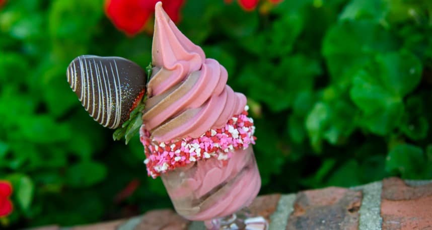Strawberry soft serve served in a glass at Disney Parks, crowned with a chocolate-dipped strawberry and pink sprinkles, set against lush greenery—the perfect Valentine’s Day treat inspired by the Disney Foodie Guide for fans seeking magical seasonal sweets.