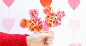 Celebrate Valentine’s Day at Disney Parks with festive Mickey Waffle Pops, decorated with hearts and served on sticks, set against a charming heart-themed backdrop—an irresistible treat for families enjoying the magic together.