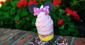 Minnie DOLE Whip Watermelon Cup featuring vibrant pink soft serve topped with a signature Minnie Mouse chocolate bow, served in a souvenir cup at Disney Springs, surrounded by colorful flowers and lush greenery.