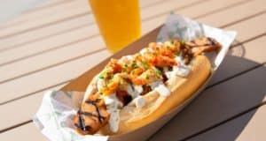 The Smoked Crawdog from Disney Springs’ Mardi Gras celebration—grilled sausage tucked in a bun with rich creamy sauce and tangy kimchi—sits ready to enjoy next to a frosty drink on a wooden table, capturing the festive spirit of the park.