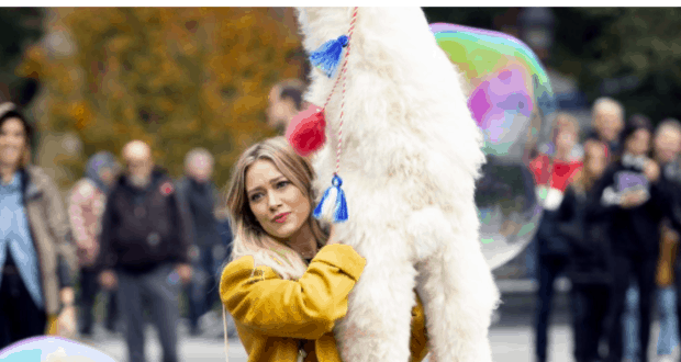 A guest in a vibrant yellow coat embraces a tall, white llama adorned with colorful tassels amidst the lively atmosphere of a festival gathering—radiating happiness that stands in vivid contrast to the buzz surrounding the cancellation of Disney’s Lizzie McGuire Revival.
