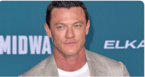 Actor in a sharp light gray suit poses in front of a teal Disney+ Pinocchio premiere backdrop, ready for the magical event celebrating Disney’s latest live-action film.