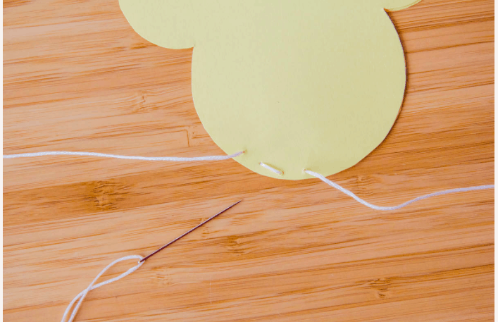 A needle and white thread rest beside a yellow paper cutout with punched holes, carefully stitched on a wooden table—an ideal craft for making Disney-themed Valentine’s Day candy pouches or enchanting Mickey-shaped treat holders to bring home some park magic.