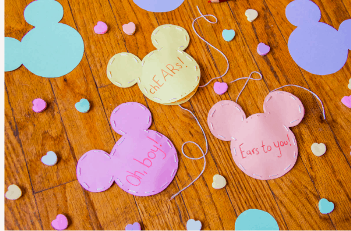 Colorful Mickey-shaped paper cutouts featuring phrases like "Oh boy!" and playful hearts are scattered across a wooden floor, perfect for bringing a bit of Disney Parks Valentine’s Day magic to your celebration.