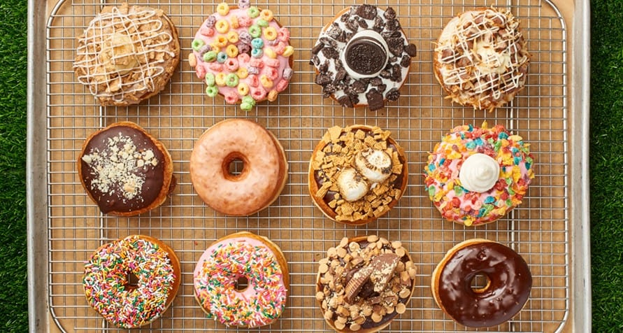 A mouthwatering lineup of twelve whimsical Everglazed Donuts sits on a cooling rack at Disney Springs, each one loaded with playful toppings like rainbow candies and pastries—an Instagram-worthy sweet treat to pair perfectly with Cold Brew during your magical visit.