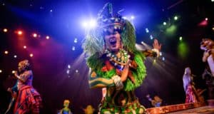 A performer in vibrant, tribal-inspired attire sings energetically on a stage illuminated by colorful lights. The lively background features other performers in equally vivid costumes, indicating a dynamic and festive performance.