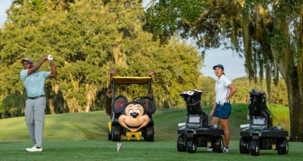 At Walt Disney World’s scenic golf course, a guest takes a swing as another enjoys the moment, while a playful golf cart adorned with Mickey Mouse’s smiling face adds classic Disney magic to the fairway experience.