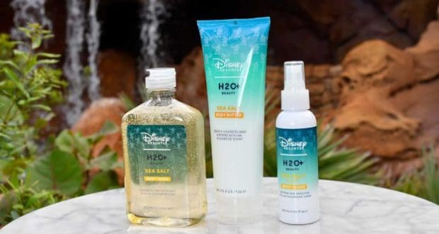 Disney Resort H2O+ bath products are displayed on a marble table outdoors, framed by lush greenery and a waterfall, immersing guests in the signature luxury of Disney Resort experiences—now available for fans to recreate Disney magic at home.