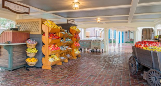 Freshly refurbished Liberty Square Market at Magic Kingdom showcases wooden carts and shelves brimming with chips, bananas, and oranges on a classic brick floor, all set beneath a welcoming covered area—perfect for grabbing a quick snack during your Disney adventure.