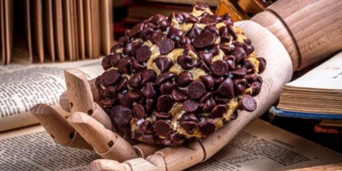 A wooden hand holding a chocolate chip cookie, surrounded by open books.