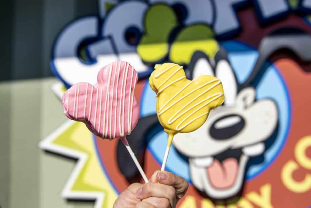 Colorful new Mickey Mouse-shaped treats in pink and yellow are displayed on sticks against the playful backdrop of a Goofy sign at Disney Springs, perfect for Disney fans looking to try the latest snacks.