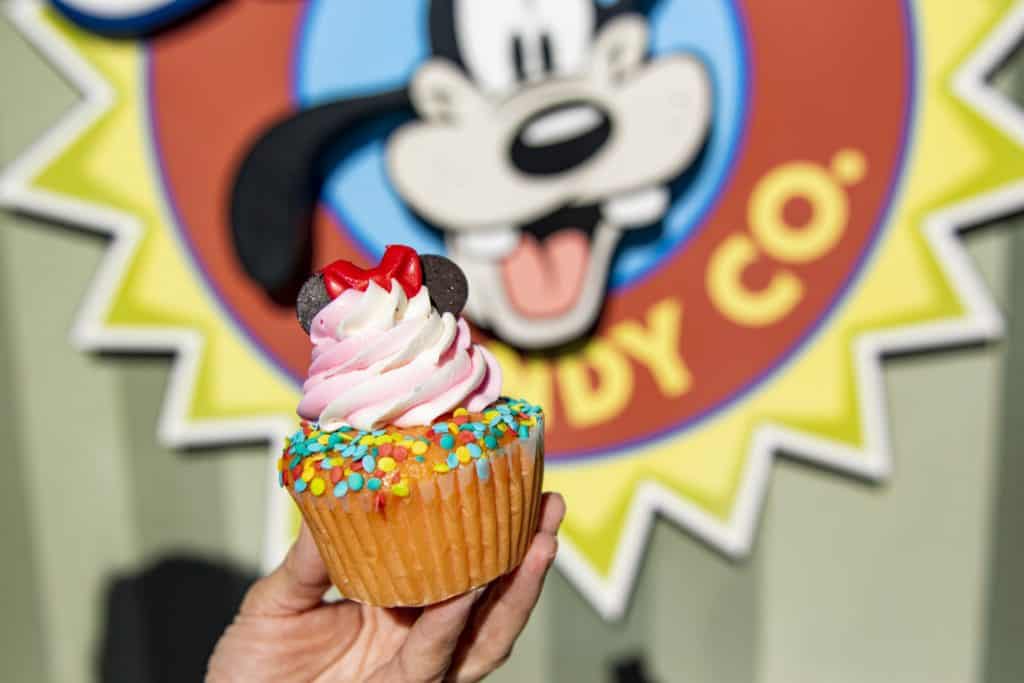A delicious cupcake topped with colorful sprinkles and iconic Minnie Mouse ears, one of the latest sweet creations at Goofy Candy Co. in Disney Springs, held up with the vibrant shop sign shining in the background—a must-try treat for Disney fans!.