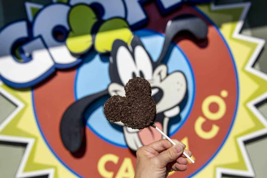 Indulge in the magic of Disney Springs with a Mickey-shaped cookie held up in front of the vibrant Goofy’s Candy Company sign—one of the latest must-try treats for Disney fans.