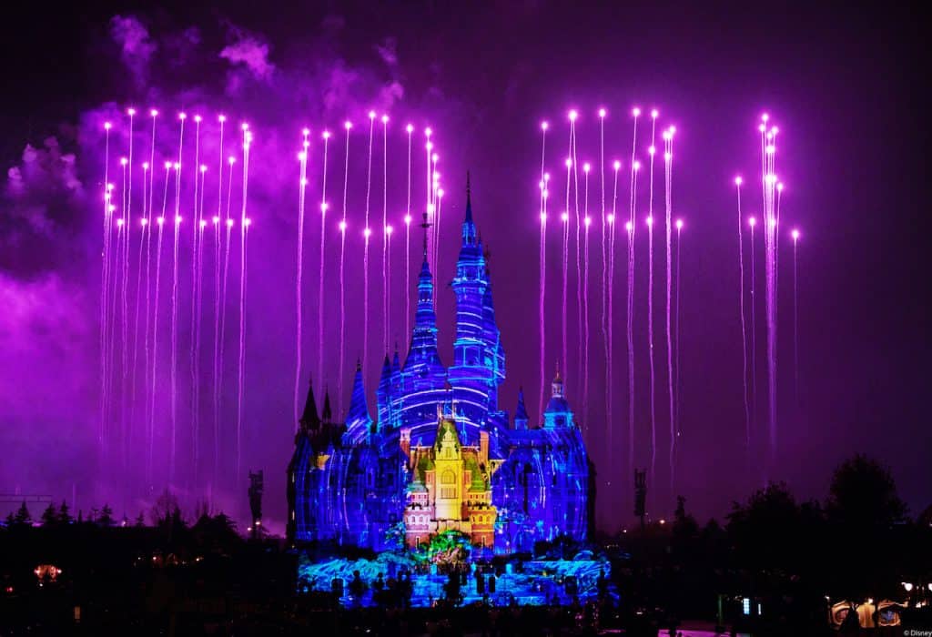 Shanghai Disneyland's Enchanted Storybook Castle illuminated against the night sky, with vibrant purple fireworks spelling out "2021" overhead to celebrate the resort’s Fifth Anniversary; the Shanghai Disney Resort logo sparkles amid a festive atmosphere.