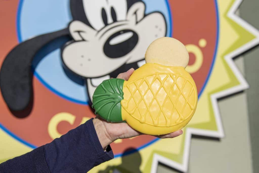 A guest shows off a Mickey-shaped pineapple cookie decorated with yellow and green icing in front of a playful Pluto sign, highlighting one of the latest snack offerings at Disney Springs.