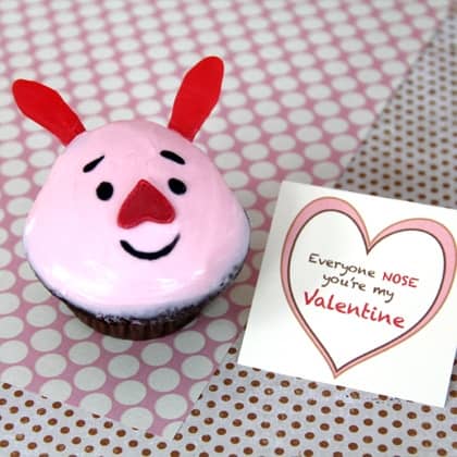 Pink Piglet Cupcake styled with a cheerful pink face, red heart-shaped nose, and matching ears sits by a Valentine card edged in hearts—this whimsical treat from our Valentine’s Day bakery collection brings sweet theme park fun straight to your celebration.