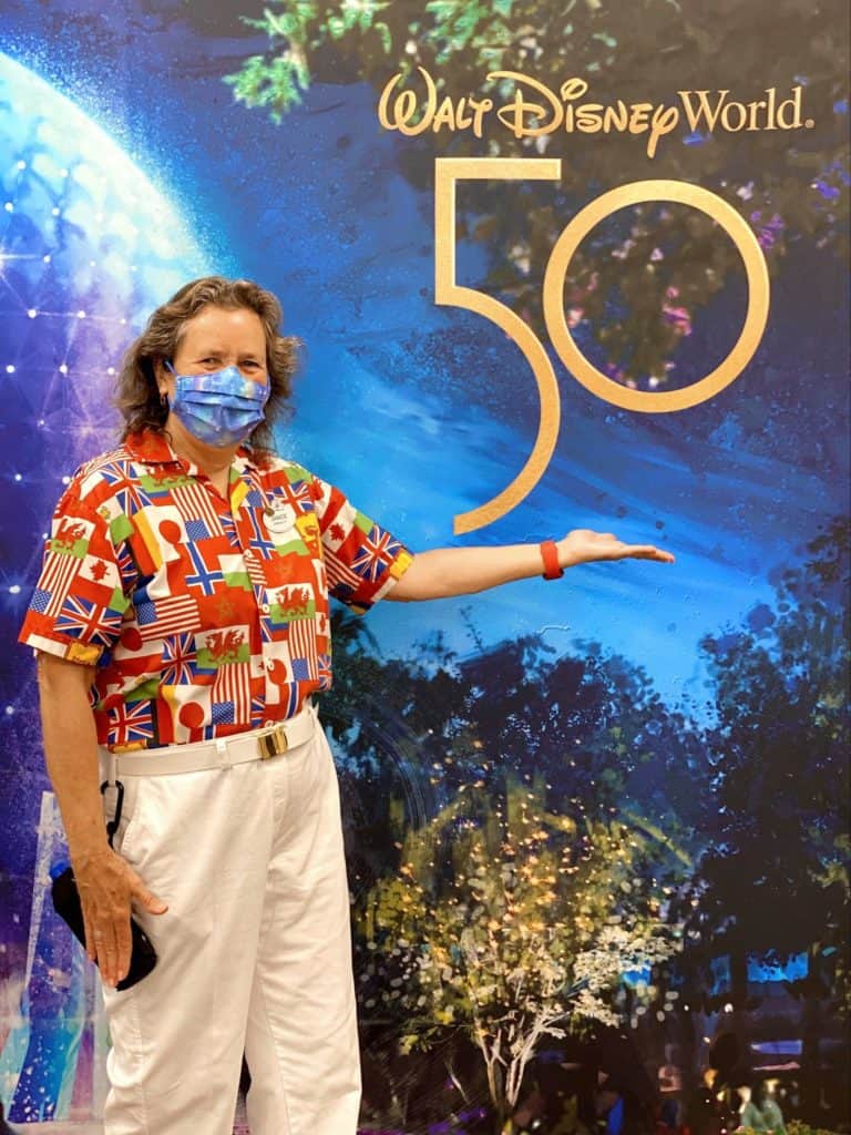 A Disney fan sporting a vibrant shirt and face mask joins Cast Members in front of the Walt Disney World 50th Anniversary sign, celebrating the park’s magical milestone together.