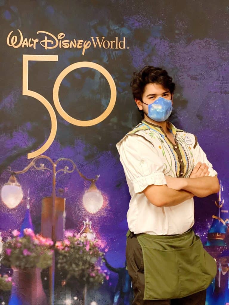 Mickey Mouse, wearing a festive mask, poses with Cast Members in front of the Walt Disney World 50th Anniversary backdrop, celebrating the magic together.