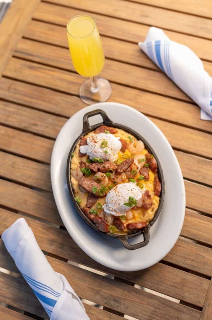 A skillet of cheesy hash crowned with poached eggs, paired with a sparkling mimosa on a rustic wooden table at The Boathouse in Disney Springs.