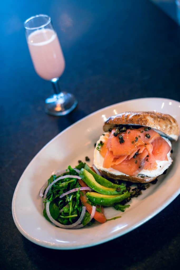 A brunch spread featuring a classic bagel topped with smoked salmon and cream cheese, an avocado salad, and a vibrant pink drink in a wine glass—setting the stage for an exciting day enjoying dueling piano shows at Jellyrolls or taking in scenic waterfront views at The Boathouse on Disney’s BoardWalk.