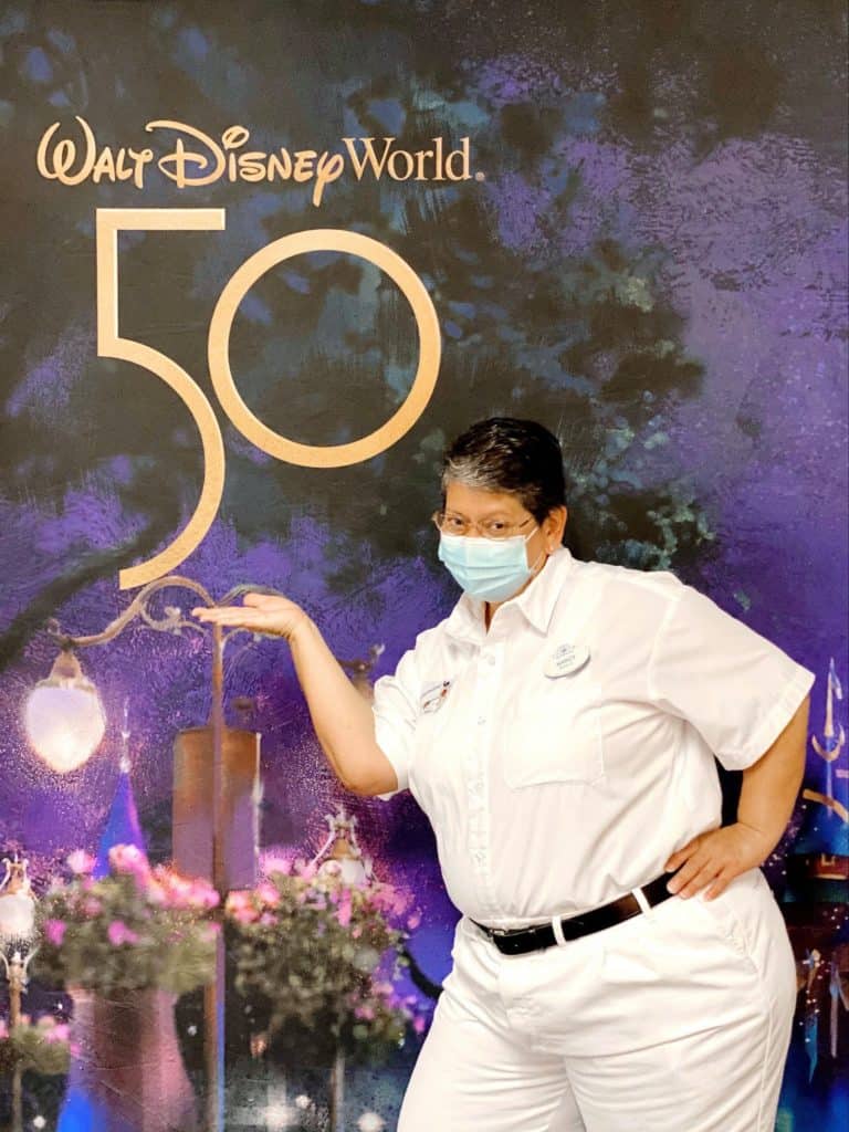 A dedicated Cast Member in a crisp white uniform and face mask stands proudly before the dazzling Walt Disney World 50th Anniversary backdrop, highlighting the magic and teamwork behind this unforgettable celebration.