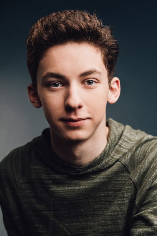 A high-spirited young guest with short brown hair in a green hoodie faces the camera against a simple backdrop, channeling iconic Season Two High School Musical vibes that infuse the scene with the excitement and freshness new visitors bring to a theme park.