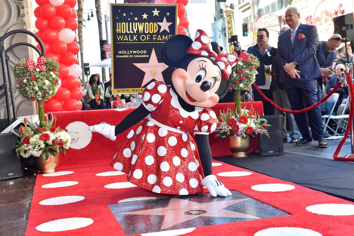 Minnie Mouse, dressed in her iconic red polka-dot dress, beams beside her Hollywood Walk of Fame star, surrounded by vibrant flowers—a magical scene that highlights the timeless appeal and Disney history celebrated by fans everywhere.