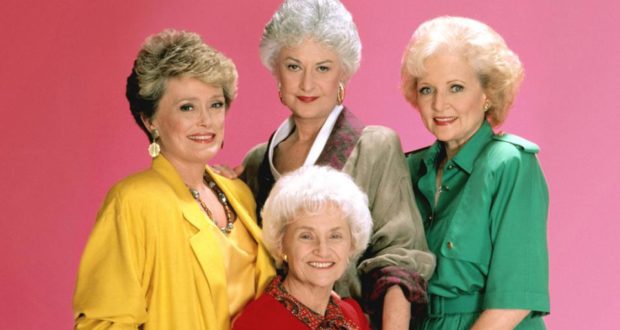 The beloved cast of The Golden Girls beams in vibrant, themed outfits against a playful pink backdrop, celebrating their timeless comedy as the show streams on Disney+.