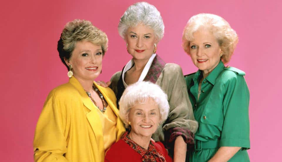 The beloved cast of The Golden Girls beams in vibrant, themed outfits against a playful pink backdrop, celebrating their timeless comedy as the show streams on Disney+.