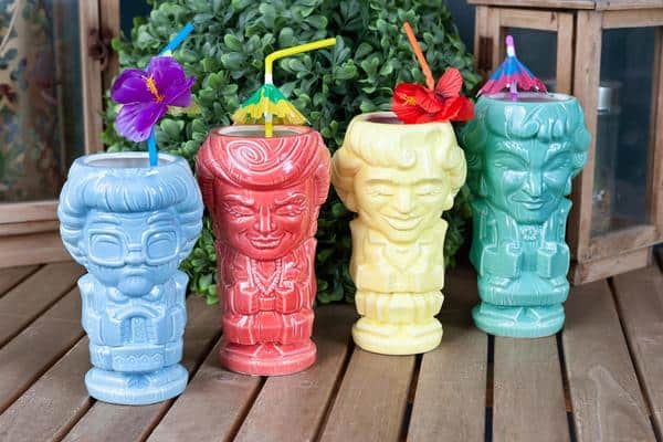 A set of four vibrant Geeki Tikis mugs themed after The Golden Girls, each topped with fun straws and decorative accents, displayed on a wooden table surrounded by lush greenery—an eye-catching collectible for fans gearing up for a Disney+ binge or bringing theme park flair to their at-home entertainment.
