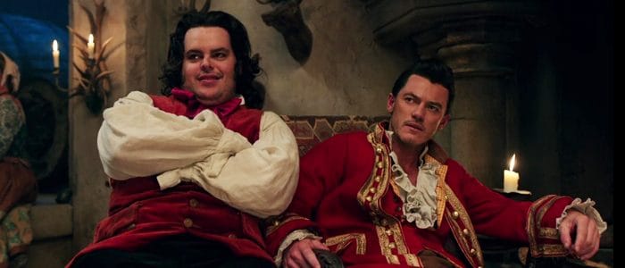 Gaston and LeFou, clad in striking red costumes, sit together inside the Disney park venue—Gaston with arms confidently crossed, LeFou leaning back—all while channeling the irresistible on-stage rapport of their iconic Luke Evans series counterparts.