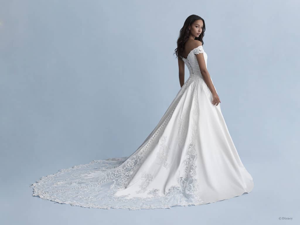 A Disney Fairy Tale Weddings bride wears a magical off-shoulder white gown with intricate lace details, standing dreamily against a light blue backdrop inspired by classic Disney elegance.
