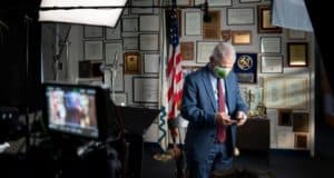 The Grinch, decked out in a suit, checks his phone in a festive office lined with awards, while professional cameras stand by—offering fans an exclusive backstage peek at Universal Studios' holiday magic.