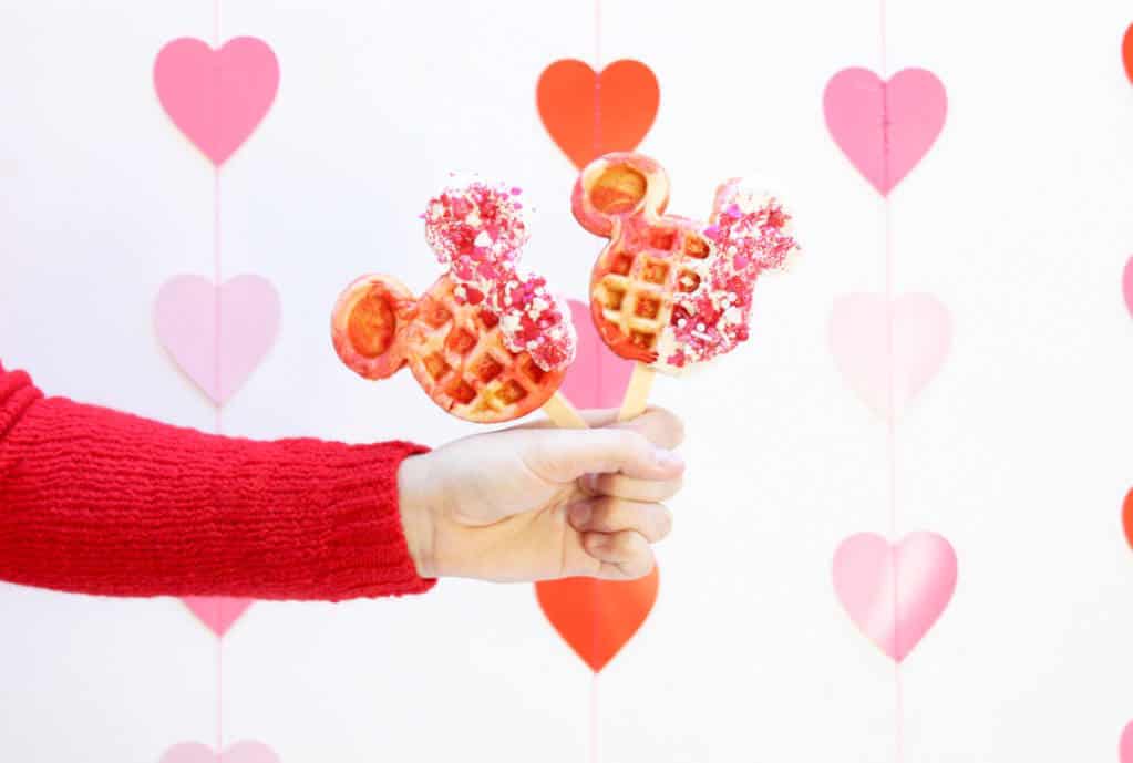 Celebrate Valentine’s Day at Disney Parks with two heart-decorated Mickey Waffle Pops held up against a festive pink and red heart backdrop—making for a magical family treat during your visit.