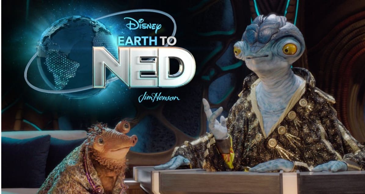 Ned and Cornelius, the alien stars of "Earth to Ned," sit in shimmering costumes at their space-age talk show desk beneath a glowing show logo, inviting Disney+ fans into an out-of-this-world late-night experience straight from the Disney universe.