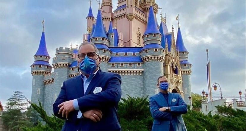 Disney World guests in festive attire and masks pose in front of the iconic Cinderella Castle, illuminated in blue and pink for the 50th Anniversary celebration at Magic Kingdom.