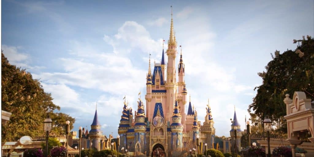 Cinderella Castle, adorned with vibrant blue and gold accents, stands majestically amid lush trees and glowing lampposts under a bright blue sky, serving as the dazzling focal point for Walt Disney World’s 50th Anniversary Park Icon Transformation.