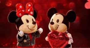 Mickey and Minnie Mouse plush toys, with their iconic big ears, stand side by side on a shimmering red background, with Minnie holding a heart—festive Disney Valentine’s collectibles that capture the magic of your favorite theme park romance.