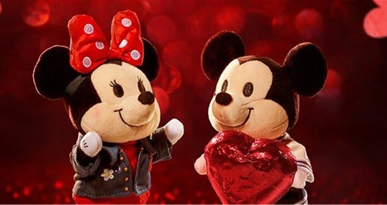 Mickey and Minnie Mouse plush toys, with their iconic big ears, stand side by side on a shimmering red background, with Minnie holding a heart—festive Disney Valentine’s collectibles that capture the magic of your favorite theme park romance.