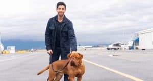On an airport runway, a man in a dark coat grins as he stands beside his loyal brown dog, capturing the heartwarming partnership of Turner & Hooch fans have come to love, now streaming on Disney+.