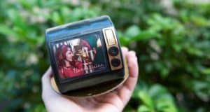 A guest holds a retro television featuring a smiling woman inspired by WandaVision, with lush theme park landscaping softly blurred in the background.