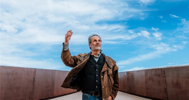 Disney Imagineer Joe Rohde, wearing his signature brown jacket, stands on a walkway beneath a clear blue sky, passionately gesturing as he shares imaginative ideas with fans outdoors.