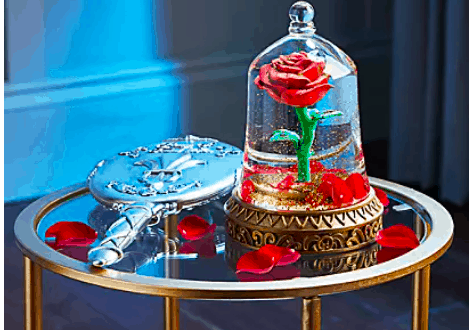 The iconic enchanted rose from Beauty and the Beast glows inside a glass dome on a round display table, surrounded by scattered petals and a sparkling silver hand mirror, evoking the magic and wonder of Disney Parks’ fairytale attractions.