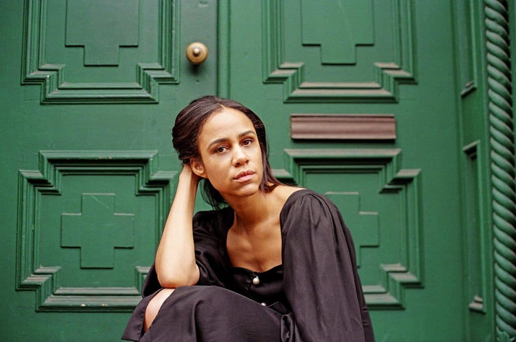 A stylish woman in a sleek black dress sits poised before the iconic green entryway reminiscent of classic movie sets, gazing at the camera with the same composed intensity as Zawe Ashton’s villainous character from Captain Marvel 2—ready for her star moment in a cinematic theme park adventure.
